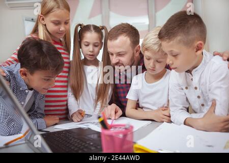 Mature male teacher enjoying working with kids at primary school, helping them with a project. Friendly teacher explaining tasks to his little student Stock Photo