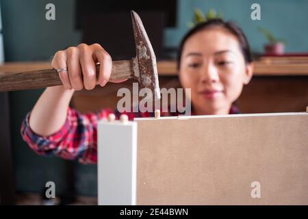 Focused ethnic female using hammer for nailing wooden dowels while assembling new furniture in apartment Stock Photo