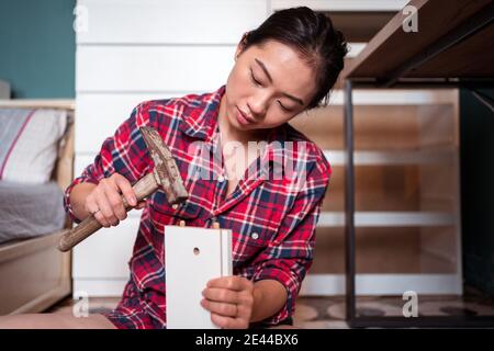 Focused ethnic female using hammer for nailing wooden dowels while assembling new furniture in apartment Stock Photo