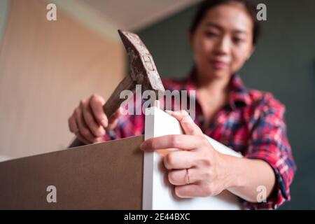 Focused ethnic female using hammer for nailing wooden dowels while assembling new furniture in apartment Stock Photo