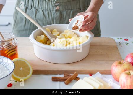 Unrecognizable person adding sugar to cut apples in baking pan near flour with lemon and butter with cinnamon during crumble preparation at home Stock Photo