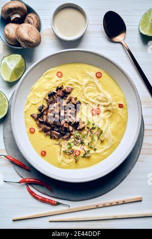 Top view of bowl with appetizing cream soup with noodles and chopped mushrooms arranged on table with chopsticks and fresh ingredients in Asian restau Stock Photo