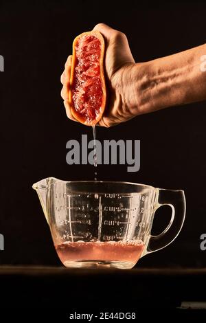 Unrecognizable crop person squeezing ripe grapefruit juice in glass measuring jug placed on table on black background Stock Photo