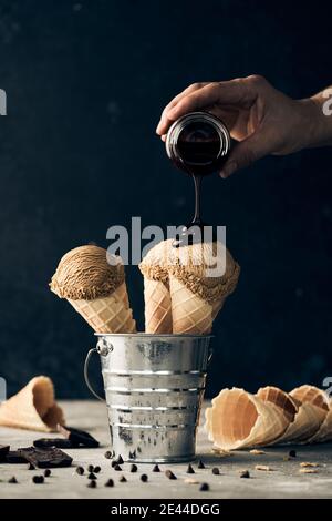 Crop anonymous person adding chocolate topping on scoops of ice cream in waffle cones placed in bucket on table on dark background Stock Photo