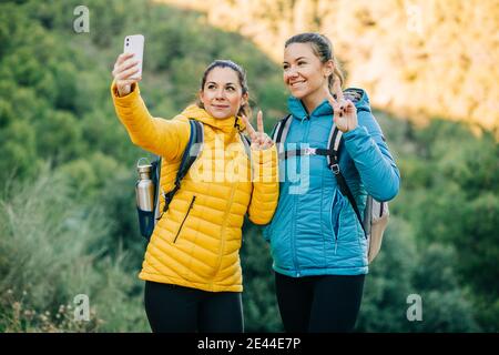 Delighted female hikers taking selfie on smartphone and showing two fingers gesture while standing in nature during trekking Stock Photo