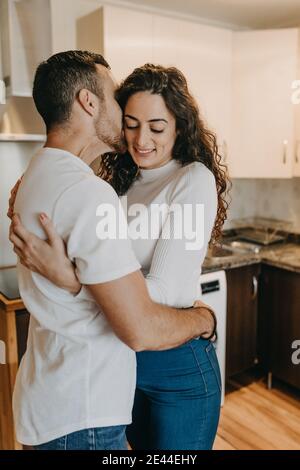 Side view of content young couple standing in modern kitchen at home and hugging while smiling and kissing Stock Photo