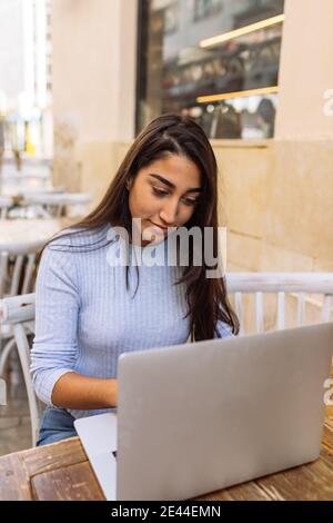Young ethnic female freelancer sitting at table and working on remote project on laptop while spending time in outdoor cafe Stock Photo