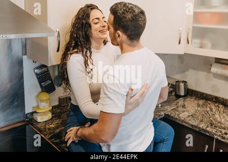 Smiling Woman sitting on counter in kitchen and embracing boyfriend while looking at each other Stock Photo