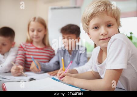 Charming little blond haired girl looking to the camera during art class, copy space. Adorable schoolgirl enjoying drawing at elementary school lesson Stock Photo