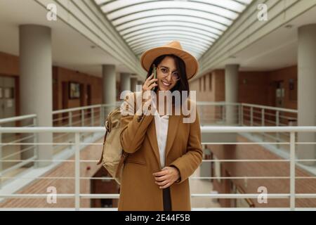Smiling female student in smart casual outfit standing in university hallway and messaging on mobile phone Stock Photo