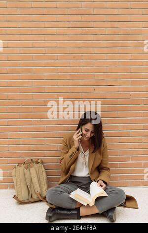 Delighted female student talking on smartphone and reading book while sitting on floor in university corridor against brick wall Stock Photo