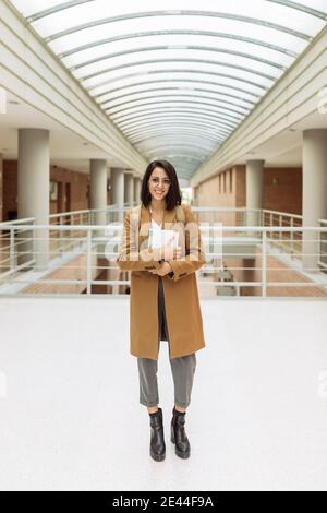Cheerful ethnic female student with backpack and pile of books standing in hallway of university building and looking at camera Stock Photo