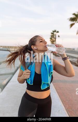 Young fit female jogger with towel on neck drinking water from bottle while having break after running on urban waterfront Stock Photo