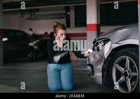 Upset young lady in casual clothes sitting on knees and touching damaged car headlights after accident in parking space Stock Photo