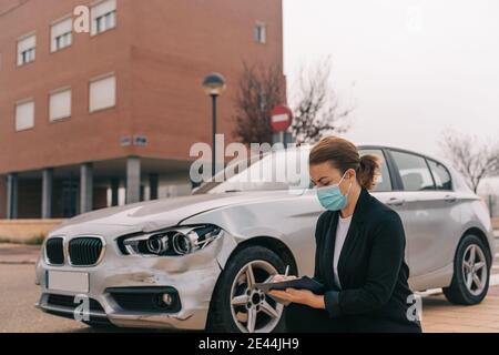 Side view of unrecognizable female insurance agent in formal outfit and protective mask writing protocol near damaged car parked on street after accid Stock Photo