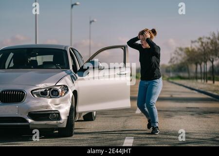 Full body of young stressed female driver in casual clothes touching head while standing on asphalt road and looking at damaged bonnet with opened mou Stock Photo