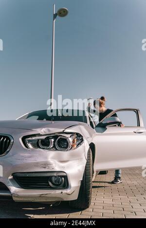 Side view of young female driver getting in modern car parked on paved road after accident against cloudless blue sky Stock Photo
