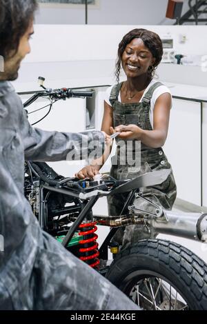 Content diverse male and female mechanics sharing tools while fixing modern custom motorbike in garage Stock Photo