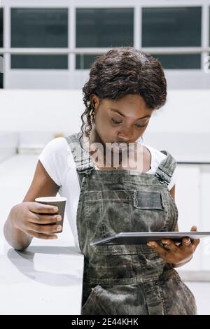Serious African American female technician in uniform using tablet and drinking coffee in workshop Stock Photo