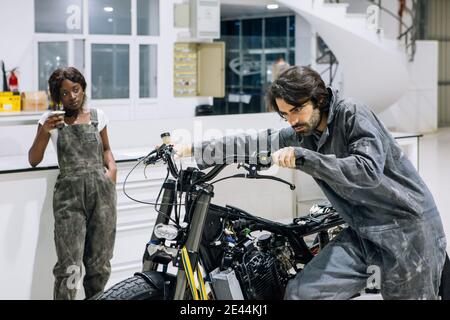 Male technician examining custom motorbike while working in garage with black female colleague having break with cup of coffee during work Stock Photo