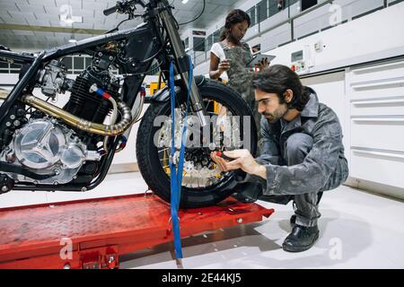 Busy African American female mechanic using tablet and drinking coffee while male technician fixing motorcycle in garage Stock Photo