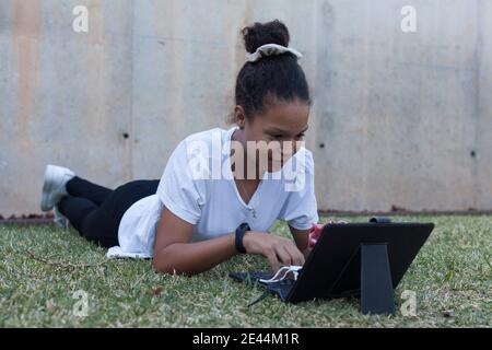 Full body of focused young African American female student in white t shirt lying on grassy lawn and typing on keyboard of modern tablet during remote Stock Photo
