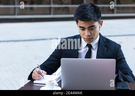 Focused Asian businessman in earbuds taking notes in notepad while sitting at table in cafe with laptop and planning business project Stock Photo