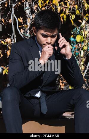 Focused Asian male entrepreneur in classy outfit sitting on bench in garden and speaking on smartphone while frowning and looking down Stock Photo