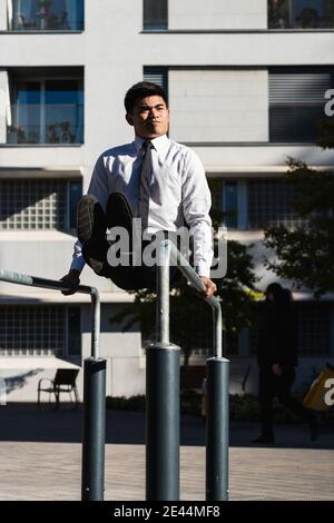 Asian male entrepreneur in formal outfit balancing on parallel bars while doing abs exercises and grimacing Stock Photo