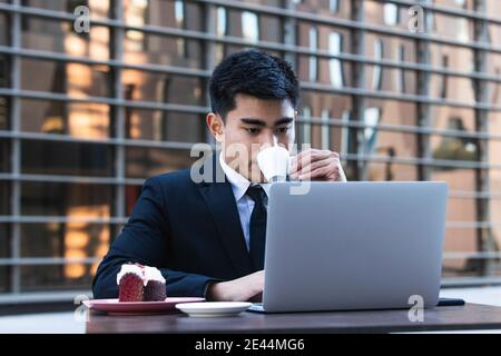 Asian male entrepreneur in classy suit sitting at table with laptop in cafe and eating sweet cake while having coffee break during work Stock Photo