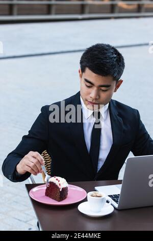 Asian male entrepreneur in classy suit sitting at table with laptop in cafe and eating sweet cake while having coffee break during work Stock Photo