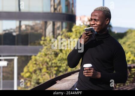 Smiling young black male in casual clothes sending voice message on smartphone while standing on terrace of modern building with cup of takeaway coffe Stock Photo