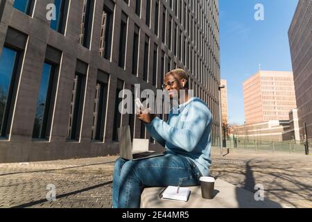 Side view of positive young black male freelancer trendy outfit and TWS earphones smiling and messaging on smartphone while working remotely on laptop Stock Photo
