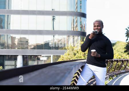 Confident young African American guy in stylish outfit using voice assistant while talking on mobile phone resting on building terrace with cup of tak Stock Photo