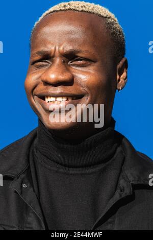 Young African American man with blond hair in black turtleneck standing against bright blue background on sunny day and looking away pensively Stock Photo