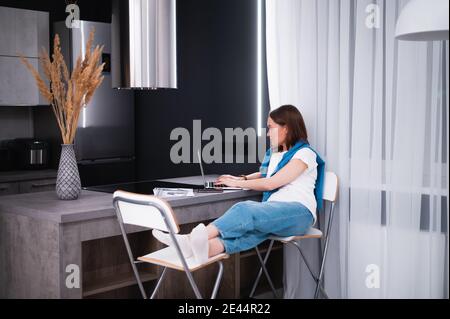 Young woman working remotly from her dining table. Home kitchen in the background. Stock Photo
