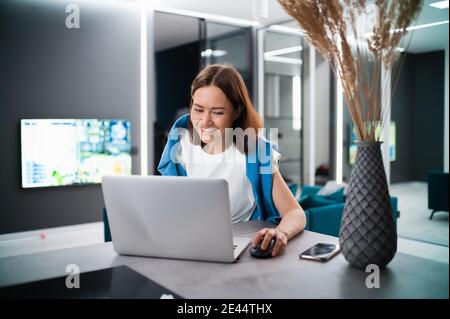 Smiling woman sitting in the kitchen and using the laptop. Working from home in quarantine lockdown. Social distancing Self Isolation. Stock Photo