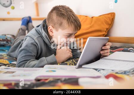 Side view of smiling teenage boy lying on bed and talking to classmates via tablet during online lesson at home Stock Photo