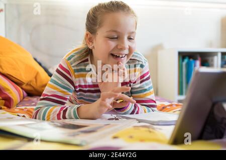 Happy schoolgirl lying on bed with tablet and having video call with classmates during online lesson from home Stock Photo