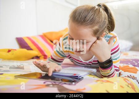 Happy schoolgirl lying on bed with tablet and having video call with classmates during online lesson from home Stock Photo