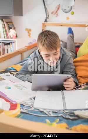 Smiling teenage boy lying on bed and talking to classmates via tablet during online lesson at home Stock Photo