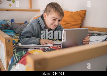 Smiling teenage boy lying on bed and talking to classmates via tablet during online lesson at home Stock Photo