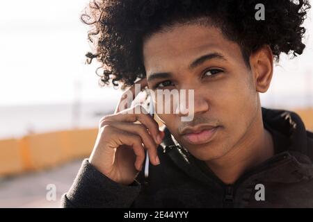 Serious African American male teenager in ripped jeans talking on smartphone while standing on street looking at camera Stock Photo