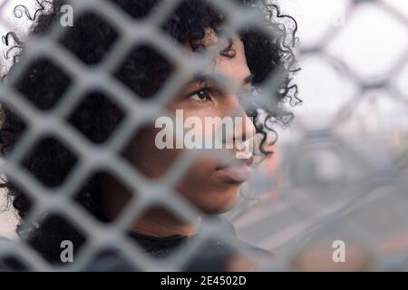 Side view of thoughtful teen African American male with curly hair looking away while standing behind metal chain link fence on urban street Stock Photo