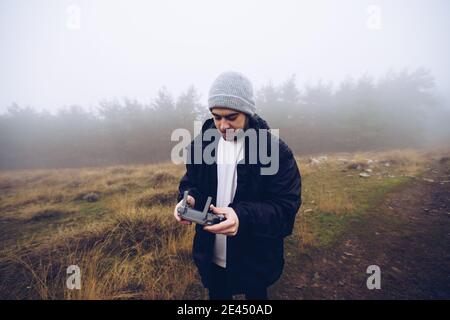 Male operating drone with remote controller while standing on path in foggy field in autumn Stock Photo