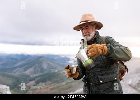 Elderly male hiker in outerwear standing in mountains in winter and pouring hot coffee from geyser coffeemaker in cup during vacation in Caceres Stock Photo
