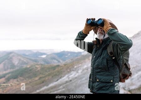 Senior male traveler in outerwear looking through binoculars while standing in highland terrain and admiring picturesque landscape during winter holid Stock Photo