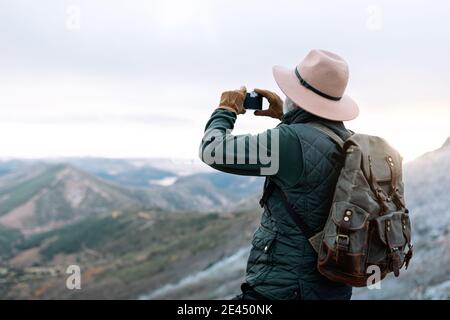 Side view of unrecognizable senior male explorer taking photo of mountainous landscape while using smartphone and enjoying vacation in Caceres Stock Photo