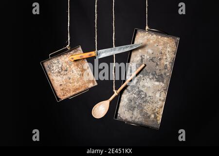 Weathered rustic metal baking sheets with knife and wooden spoon hanging on ropes against black background Stock Photo
