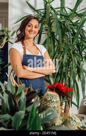 Smiling female gardener in apron standing with crossed arms in flower store with assorted potted plants and looking at camera Stock Photo